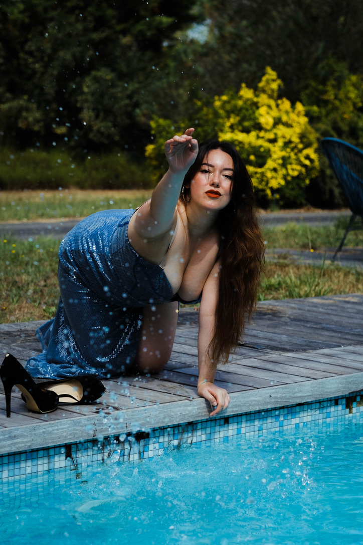 Kneeled woman wearing a green shining dress splashing water from the pool.